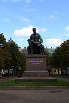 A statue of a tall man with a long beard and glasses, seated, a large musical score open in his lap