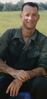 A man is at the center of the image smiling into the camera. He is sitting on a blue crate and has his hands resting on his legs.