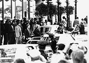 A chaotic scene of motorcade vehicles surrounded by crowd of people including police and press