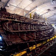 A diagonal section of a wooden ship resting upright on a yellow steel frame inside a building with bare, drab walls and sprinkler systems in the ceiling