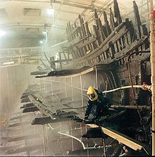 A diagonal section of a wooden ship seen from the stern while being sprayed with a clear liquid from a built-in sprinkler system; on the remnants of the main deck there is a person in a black, full-body protective plastic suit with a yellow helmet