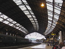  A railway station with curved platforms under an arched iron framed roof with roof-lights. A passenger train stands at the platform on the right and on the left passengers waiting for a train.