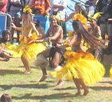 Two women in long flowing yellow skirts either side of a man in a short black skirt mid dance