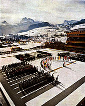 An open air arena with crowds in the stands and people on the playing field surrounding a rostrum