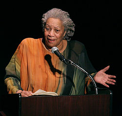 Photograph of Toni Morrison, showing her in an orange and green dress with her gray hair pulled back. She is speaking at a podium and gesturing.