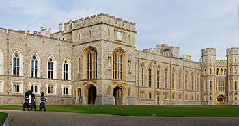 A photography of a grey Gothic building stretching across the picture; the nearest parts have windows in a white stone, the furthest parts in a yellow stone. Three soldiers in grey uniforms and black hats are marching past the building.