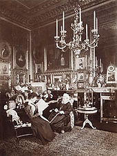 A black and white photograph of an elderly Victoria sat alongside a younger woman (Beatrice) reading a newspaper. The room is ornately decorated, with a number of photographs, paintings and a large chandelier hanging from the ceiling.