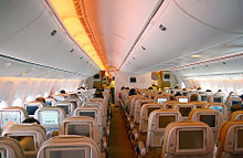 Airliner cabin. Rows of seats arranged between two aisles. Each seatback has a monitor; light shines from the sidewalls and overhead bins.