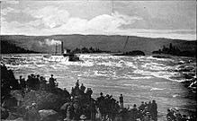 A river boat with more than a dozen windows along its visible side runs a set of rapids on a very large river. Smoke or steam rises from its smokestack and flows behind the boat parallel to the water. In the foreground, a crowd of 50 or people watch the boat from the rocky shore.