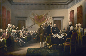 About 50 men, most of them seated, are in a large meeting room. Most are focused on the five men standing in the center of the room. The tallest of the five is laying a document on a table.