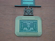 A stone tablet, inscribed with the image of a football pitch and several names. It is surrounded by a stone border in the shape of a football stadium. Above the tablet is a wooden carving of two men holding a large wreath.