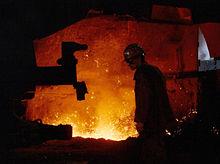 An elderly worker in a helmet is facing his side to the viewer in an industrial hall. The hall is dark but is illuminated yellow glowing splashes of a melted substance.