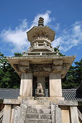 A stone pagoda with elaborated tiers, a small lion status, and stairs. Blue skies and a roof of a building and trees are shown on the background