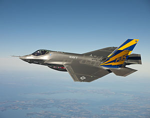 Gray fighter aircraft flying in a clear blue sky with sea coast below.