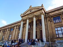 The façade of a masonry building, with four Greek adorning its entrance, under a clear blue sky