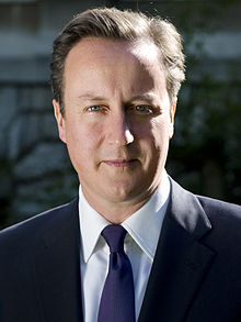 A man, clean shaven, with short straight dark brown swept back hair wearing a suit jacket, white shirt and blue tie