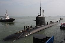 A Scorpène class submarine at dock, half out of the water. People on top are mooring it, and a boat can be seen in the background