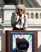 A blond woman speaking on a kiosk. She wears a white shirt and black glasses. Behind her, the balcony of a building is visible.